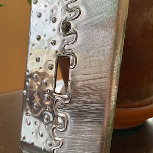 May include: A silver metal light switch cover with a textured surface and a decorative border. The cover has a raised, circular pattern and a wavy design.
