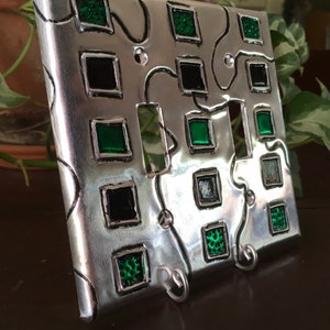 May include: A silver metal light switch cover with a decorative design featuring black and green squares. The cover has a textured surface and is accented with small metal hooks.