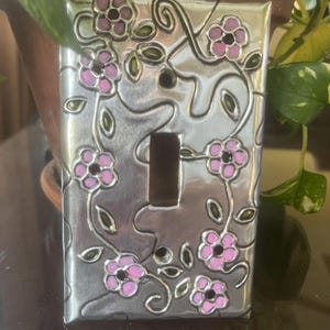 May include: A decorative light switch cover with a floral design. The silver-colored cover features pink flowers with black centers and green leaves. The flowers and leaves are arranged around the switch opening.
