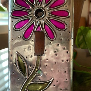 May include: A silver metal light switch cover with a pink flower and green leaves. The flower has a textured, hammered metal look.