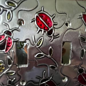 May include: Silver-colored metal light switch cover with a design of ladybugs and leaves. The ladybugs are red with black spots and outlines. The leaves are green and the background is silver.