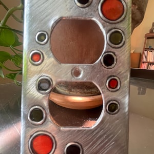 May include: A decorative, brushed metal outlet cover with two openings and several smaller, colorful, circular accents. The accents are in shades of red, green, and black. The cover is rectangular with rounded corners.