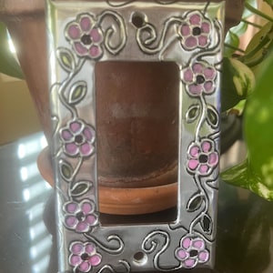 May include: A silver-colored light switch cover with a floral design. The cover features pink flowers with black centers and green leaves, connected by swirling vines. The rectangular cover is made of metal.