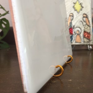 May include: White ceramic tile with a gold metal stand. The tile has a smooth, glossy finish and is blank. The stand has two heart-shaped hooks.