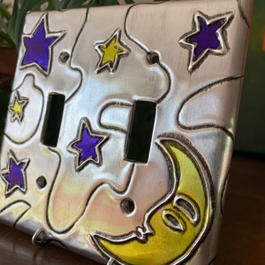 May include: A silver metal light switch cover with a yellow crescent moon and blue and yellow stars. The cover has a brushed metal finish.