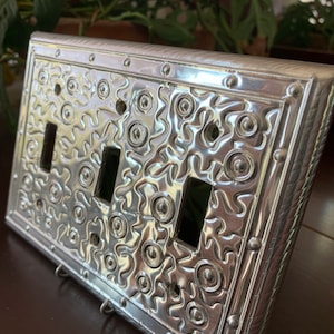May include: A silver metal light switch cover with a decorative embossed pattern. The cover has three switches and a textured border.