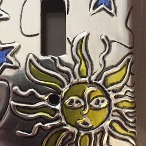 May include: A silver light switch cover with a sun, stars, and moon design. The sun is yellow with a face and the stars are blue.
