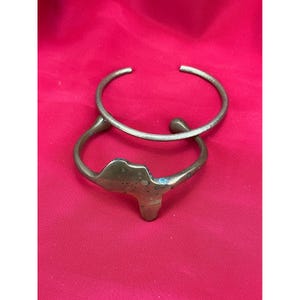 May include: Two brass-toned metal bracelets on a red background. One is a simple open cuff, and the other features a stylized map of Africa. The bracelets are slightly tarnished, showing signs of age and wear.