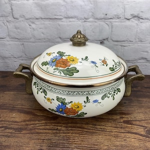 May include: A white enamel casserole dish with a lid, featuring a floral design in blue, orange, and yellow. The dish has brass-colored handles and trim, and a matching brass knob on the lid. The dish is on a wooden surface.