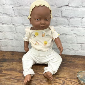 May include: A dark-skinned baby doll wearing a cream-colored bonnet, a short-sleeved onesie with colorful graphics, and white pants. The doll is sitting on a wooden surface.