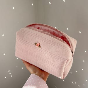 May include: A light pink rectangular cosmetic bag with a zipper closure. The bag features a delicate, textured pattern and a small, embroidered rose. The interior lining is also pink.