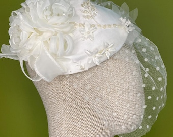 May include: A white bridal hat with a floral arrangement and a dotted veil. The hat is adorned with fabric flowers, ribbons, and small star-shaped embellishments. The veil is a sheer, white fabric with small, evenly spaced white dots.