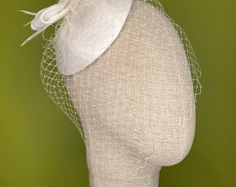 May include: A white fascinator with a bow and feather accents, and a delicate net veil. The headpiece is displayed on a mannequin head against a green background. The hat is a formal accessory.