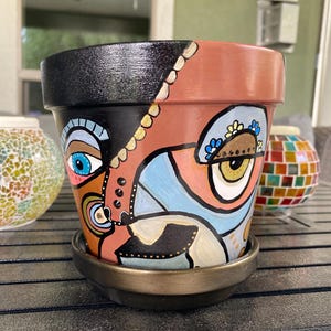 May include: A hand-painted terracotta flower pot with a whimsical face design. The pot features a black top, a brown base, and a face with blue, orange, and yellow details. It sits on a matching saucer.