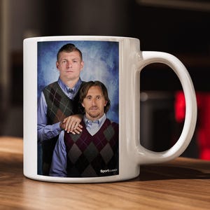May include: A white ceramic mug featuring a printed photograph of two individuals wearing light blue collared shirts and argyle sweater vests against a blue mottled backdrop. The text SportClassics is printed in white at the bottom right corner.