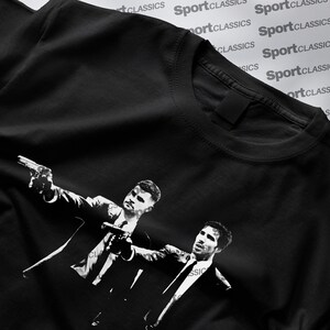 May include: Black t-shirt with a monochrome graphic of two men in suits, each holding a gun. The illustration is a stylized design. The t-shirt has a classic crew neck. The words 'SportClassics' are printed on the shirt.
