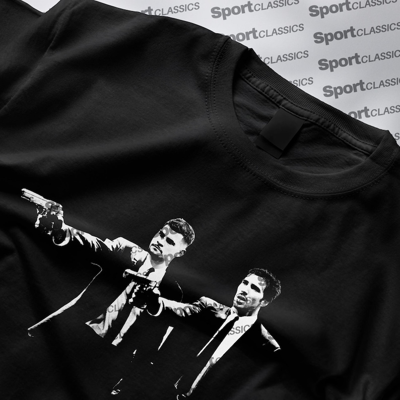 May include: Black t-shirt with a monochrome graphic of two men in suits, each holding a gun. The illustration is a stylized design. The t-shirt has a classic crew neck. The words 'SportClassics' are printed on the shirt.