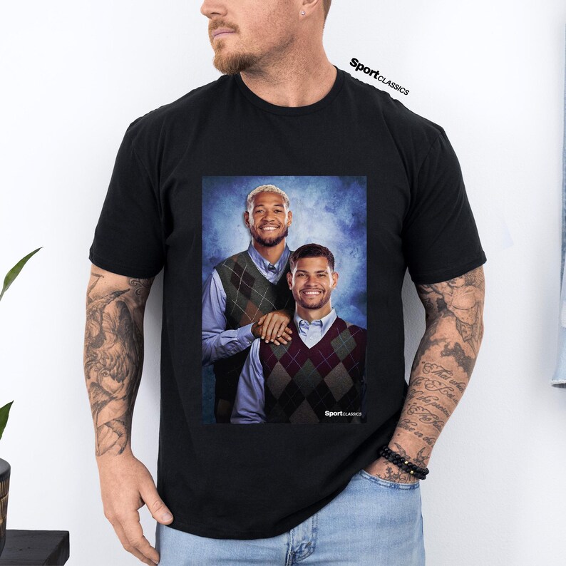 May include: Black t-shirt featuring a portrait of two smiling men wearing argyle vests over collared shirts. The image has a blue background and the text "SportClassics" is visible.