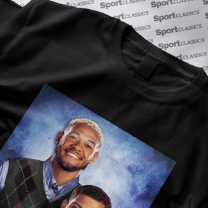 May include: Black crew neck t-shirt with a printed image of two smiling men. The image is on a blue background. The t-shirt is made of a soft material. The background has the words "SportClassics" printed on it.