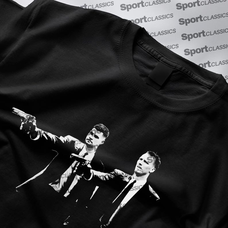Pu&ograve; includere: T-shirt nero con grafica in bianco e nero di due uomini in abito che imbracciano pistole. L'immagine &egrave; un'illustrazione stilizzata. La maglietta ha un girocollo ed &egrave; realizzata in materiale morbido.
