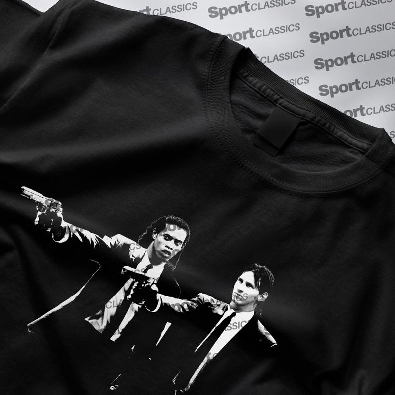 May include: Black t-shirt featuring a black and white graphic of two men in suits holding guns. The graphic is a stylized image. The shirt has a crew neck and short sleeves. The words "Sport Classics" are printed in white diagonally across the top of the shirt.