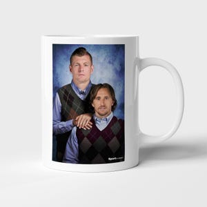 May include: A white ceramic mug featuring a printed photograph of two individuals wearing light blue collared shirts and argyle sweater vests. The individuals are posed against a mottled blue background, with the text SportClassics at the bottom.
