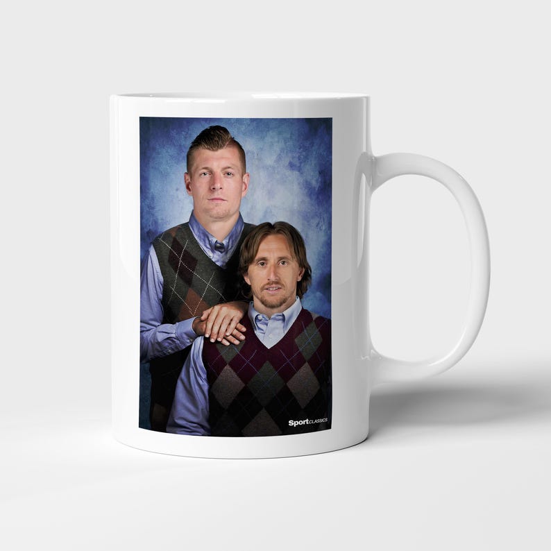 May include: A white ceramic mug featuring a printed photograph of two individuals wearing light blue collared shirts and argyle sweater vests. The individuals are posed against a mottled blue background, with the text SportClassics at the bottom.
