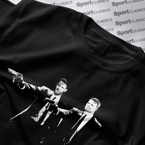 May include: Black t-shirt with a monochrome graphic of two men in suits, each holding a gun. The design is a stylized illustration. The shirt has a crew neck and short sleeves. The words "SportClassics" are printed repeatedly in the background.