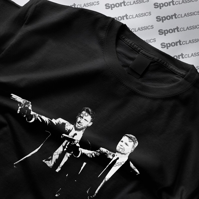 May include: Black t-shirt with a monochrome graphic of two men in suits, each holding a gun. The design is a stylized illustration. The shirt has a crew neck and short sleeves. The words "SportClassics" are printed repeatedly in the background.