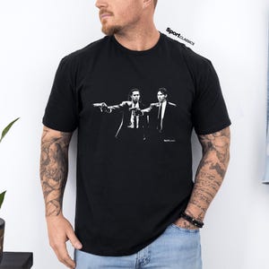 May include: Black t-shirt featuring a white graphic of two men in suits holding guns. The text "SportClassics" is printed at the top right. The image is a stylized illustration.