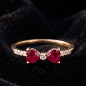 Op de afbeelding: Een gouden ring met een strikvormig ontwerp bestaande uit twee hartvormig geslepen rode edelstenen en een kleine ronde heldere edelsteen in het midden. De ringband is aan de zijkanten bezet met een reeks kleine heldere steentjes.