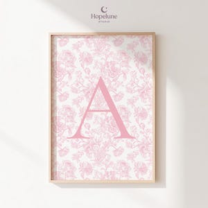 Pode incluir: Impressão artística emoldurada com um padrão floral rosa e a letra "A" rosa. A obra de arte tem uma moldura de madeira clara e está pendurada em uma parede branca. O logotipo do Hopelune Studio é visível no canto superior direito.