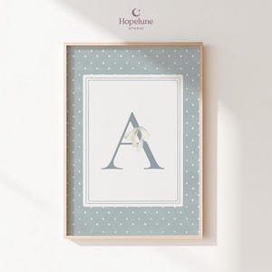 Peut inclure: Impression encadrée avec la lettre "A" en bleu clair, ornée d'un ruban blanc délicat. Le fond est bleu clair à pois blancs, le tout encadré dans un cadre en bois. Le logo "Hopelune Studio" est visible en haut à droite.