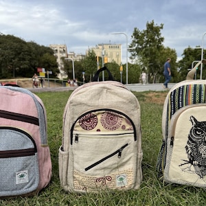 Może przedstawiać: Trzy plecaki stojące obok siebie. Lewy jest różowo-niebieski z dwiema kieszeniami na zamek. Środkowy jest beżowy w bordowy wzór kół z ukośnym zamkiem. Prawy jest kremowy z czarną grafiką sowy i pasami na górnej części oraz po bokach.