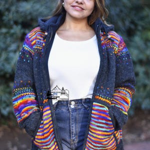 Puede incluir: Chaqueta con capucha gris oscuro con un vibrante diseño de rayas arcoíris en las mangas y la parte delantera. La chaqueta tiene cierre de cremallera y una camisa blanca debajo. La chaqueta está hecha de un material grueso y tejido.