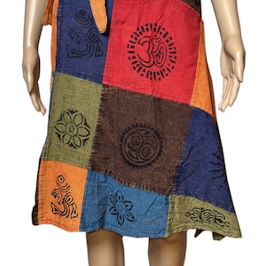 Op de afbeelding: Een knielange overslagrok met een patchworkontwerp van verschillende gekleurde stoffen vierkanten in blauw, rood, bruin, oranje en groen. De rok is versierd met zwarte bedrukte symbolen, waaronder Om-tekens, zonnen en bloemmotieven, en heeft een oranje strikceintuur.