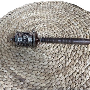 May include: A prayer wheel with a silver and dark brown barrel, featuring etched floral designs and script. The handle is dark wood. The prayer wheel is resting on a woven, circular mat made of natural fibers.
