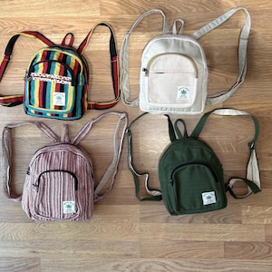 Può includere: Quattro piccoli zaini con spallacci regolabili e tasche frontali con cerniera. Uno presenta strisce verticali multicolori, uno è color crema, uno è verde oliva e uno ha un motivo a trama fine rosso e bianco.