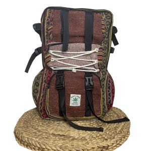 Pode incluir: Mochila multicolorida com design tecido, apresentando padrões em tons de castanho, vermelho e amarelo. Possui alças e fivelas pretas, e um cordão branco. O texto "HIMALAYA HEMP" é visível em uma pequena etiqueta.