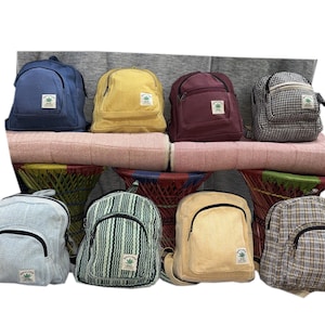 May include: A collection of eight small backpacks in various colours and patterns, including solid blue, yellow, maroon, and beige, alongside checkered and striped designs. Each bag features a front zippered pocket and a small white label with a green leaf.