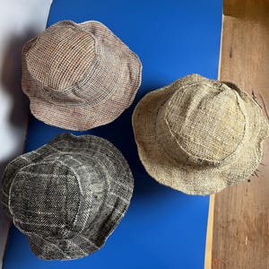 Può includere: Tre cappelli a secchiello con un aspetto intrecciato e testurizzato. Un cappello è di colore marrone chiaro, un altro è un mix multicolore di fili marroni, blu e bianchi, e il terzo è grigio antracite scuro con macchie bianche sul tessuto.
