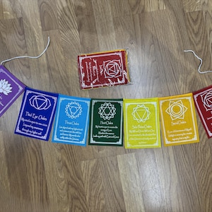 Può includere: Un set di sette bandiere chakra rettangolari nei colori viola, blu, azzurro, verde, giallo, arancione e rosso. Ogni bandiera presenta un simbolo geometrico bianco, il nome di un chakra e un'affermazione. Una bandiera rossa è ripiegata.