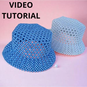 May include: Two beaded bucket hats, one blue and one silver, sit on a pink surface. The hats are made of small, round beads, creating a textured, open weave design. The words "VIDEO TUTORIAL" are in the top left corner.