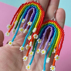May include: A pair of handmade beaded earrings in a rainbow design. The earrings feature a curved rainbow arch in red, orange, yellow, green, blue, and purple. Dangling from the arch are beaded strands with small white daisy accents. The earrings are held in a hand.