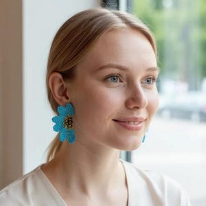 May include: A woman wearing large, turquoise beaded flower earrings. The earrings have a detailed, textured appearance with a gold-colored center. The woman is fair-skinned with blonde hair and is wearing a white top.