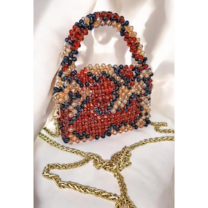 May include: A beaded handbag with a gold chain strap. The bag is covered in red, black, and tan beads, with a matching beaded handle. The gold chain is draped across a white surface.
