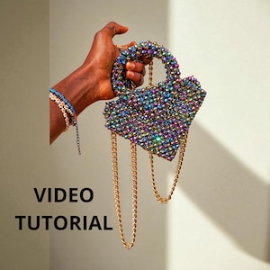 Puede incluir: Un bolso con forma de corazón adornado con cuentas con una cadena dorada. El bolso está cubierto de cuentas iridiscentes que reflejan múltiples colores. Las palabras "VIDEO TUTORIAL" están impresas en letras negras.