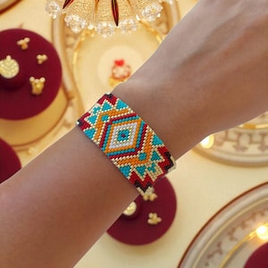 Puede incluir: Una pulsera ancha de cuentas con un patrón geométrico en rojo, naranja, turquesa y blanco. La pulsera se lleva en la muñeca, mostrando el intrincado trabajo de cuentas y los colores vibrantes. El diseño presenta formas de diamante y triángulo.