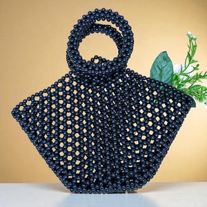 Può includere: Una borsa a mano di perline blu navy scuro con un manico circolare. La borsa è di forma quadrata con un design a griglia e un rametto di fiori bianchi e foglie verdi è infilato all'interno. La borsa è su una superficie bianca con uno sfondo dorato.