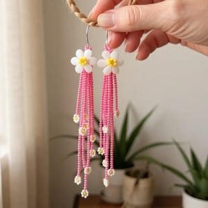 May include: A pair of handmade dangle earrings with white daisy flower tops and yellow centres. The earrings have long, pink beaded fringe with small daisy accents. The earrings are held up by a hand.